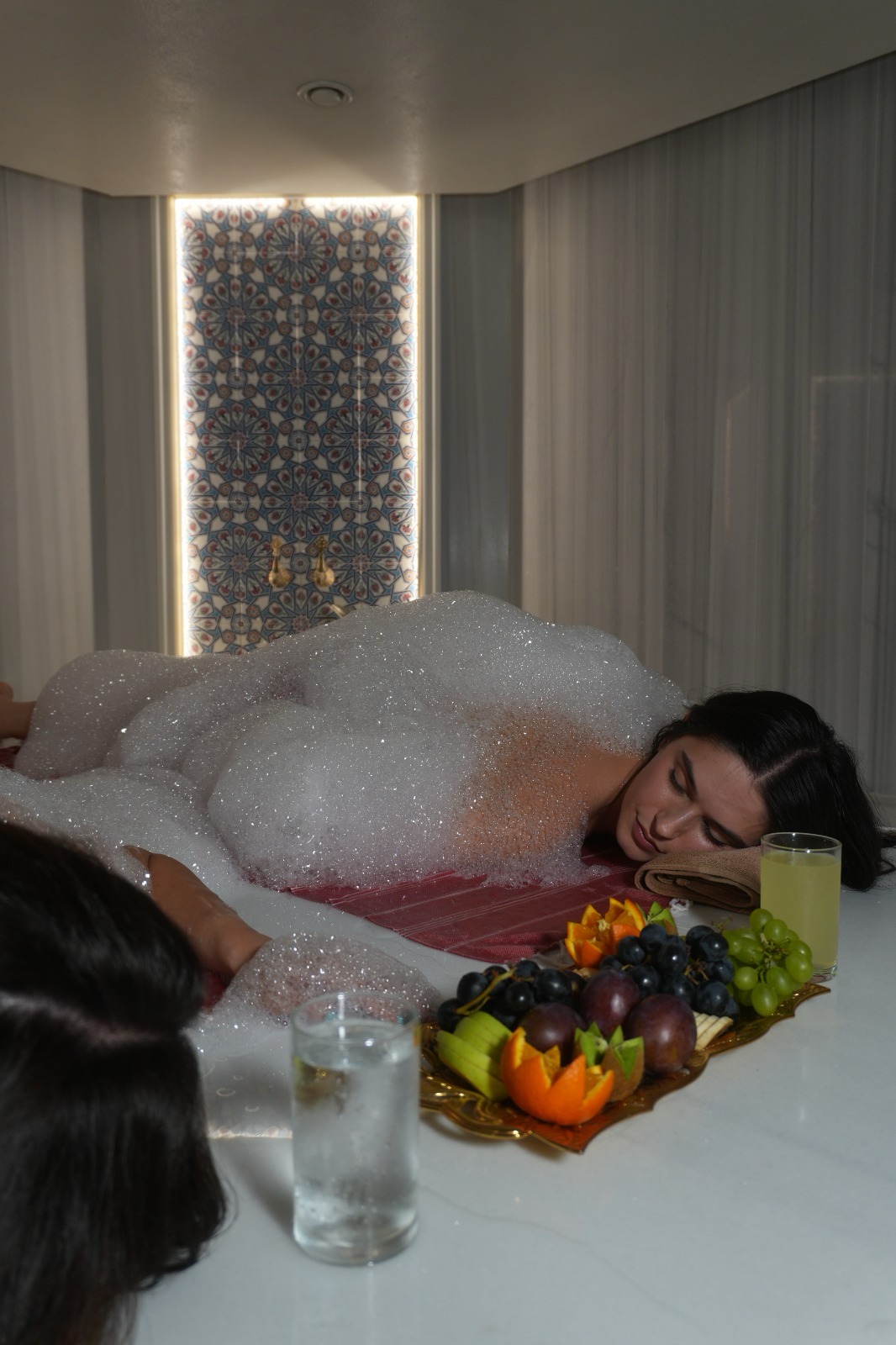 Hamam taşında sabun köpüğüne gömülmüş, gözleri kapalı uzanan bir kadın; önünde meyve tabağı ve iki bardak, arkada ışıklı çini panel.