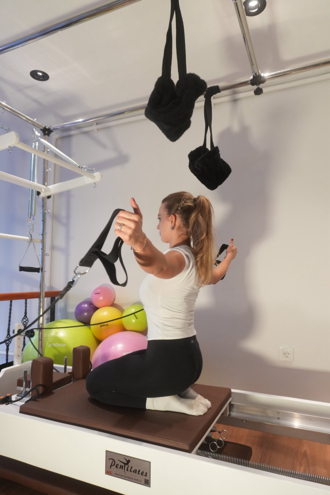 Beyaz tişörtlü, atkuyruklu bir kadın reformer üzerinde diz çökmüş, kayışları tutarak kollarını açıyor; arkada renkli pilates topları.