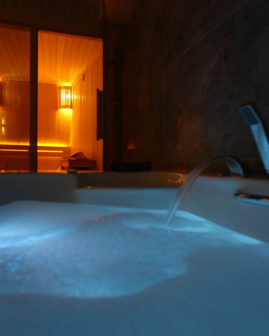 Loş jakuzi alanında mavi ışıklı su ve arkada sauna ışığı.